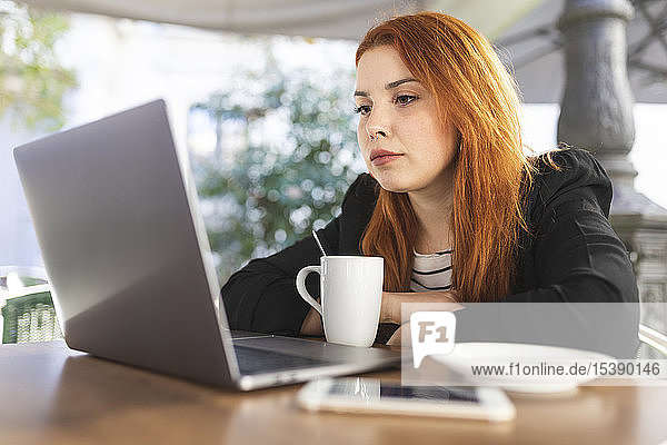 Porträt einer rothaarigen jungen Frau im Straßencafé  die auf ihren Laptop schaut