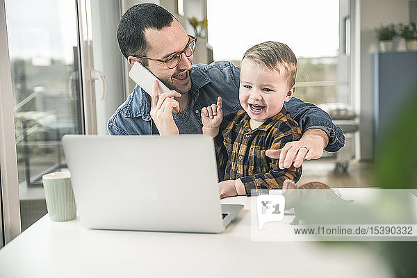 Glücklicher Vater am Handy im Heimbüro mit dem Sohn auf dem Schoß