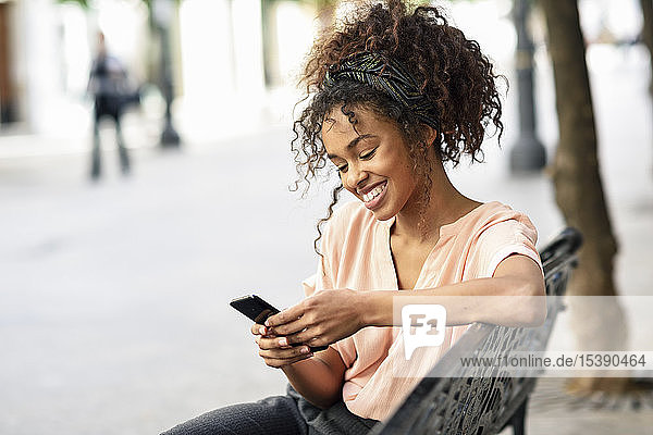 Lächelnde junge Frau sitzt auf einer Bank und benutzt ihr Handy
