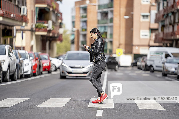 Junge Frau  die den Fußgängerüberweg überquert und ein Mobiltelefon benutzt