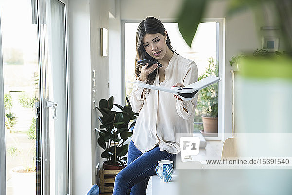 Junge Frau sitzt zu Hause am Tisch und hält Smartphone und Flugzeugmodell in der Hand