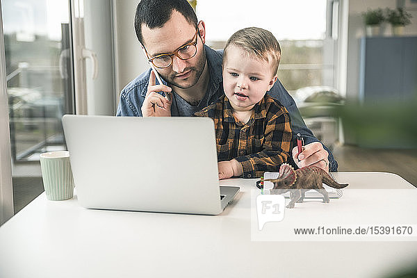 Father working at table in home office with son sitting on his lap