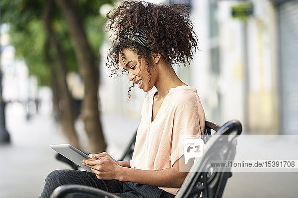 Lächelnde junge Frau sitzt auf einer Bank und benutzt ein Tablett