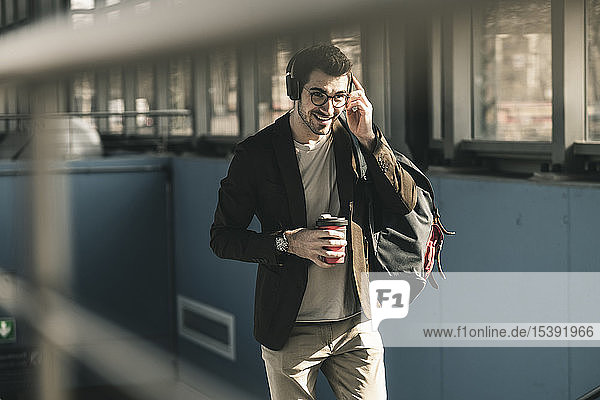 Lächelnder junger Mann mit Kopfhörern  Handy und Kaffee zum Mitnehmen auf dem Bahnhof
