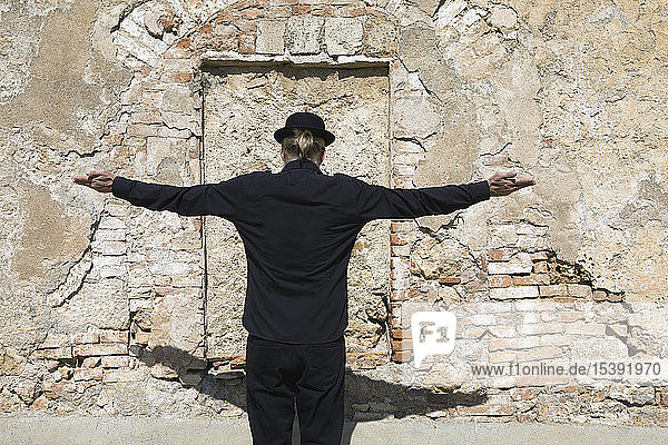Rear view of man wearing a bowler hat standing at stone wall with arms outstretched