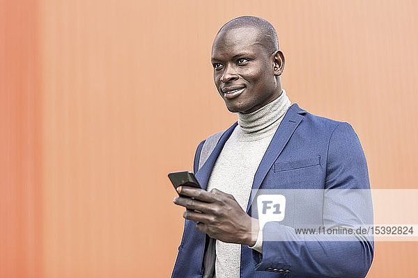 Porträt eines lächelnden Geschäftsmannes mit Smartphone vor oranger Wand