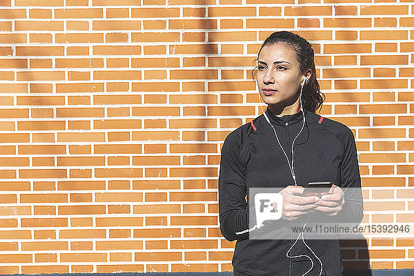 Portrait of sporty young woman with earphones and cell phone at a brick wall