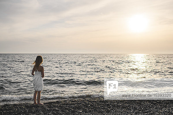 Kleines Mädchen in weißem Kleid  das den Sonnenuntergang über dem Meer betrachtet