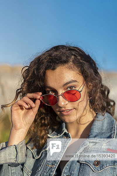 Bildnis einer jungen Frau mit lockigem braunen Haar und roter Sonnenbrille