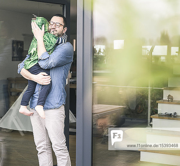 Anhänglicher Vater  der seinen Sohn in einem Kostüm an der Terrassentür zu Hause trägt