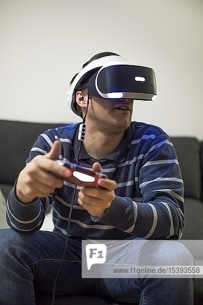 Junger Mann mit VR-Brille sitzt zu Hause auf der Couch und spielt Videospiel