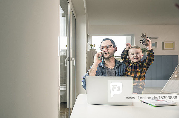 Father working at table in home office with son sitting on his lap