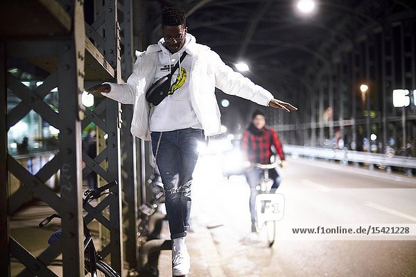 Young man balancing on metal construction of bridge Hackerbrücke Hacker bridge at night in city next to street bicycle driver in Munich Germany