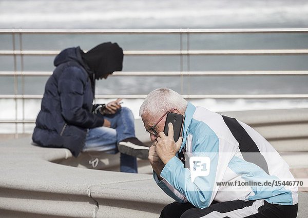 Elderly man and young man using mobile phones ovelooking city beach in Las Palmas Gran Canaria Canary Islands Spain.