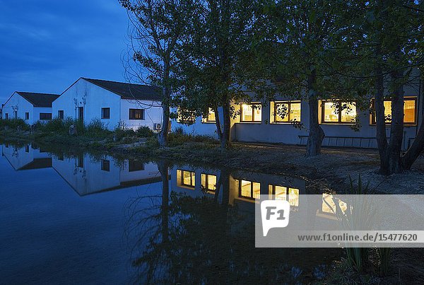The lighted restaurant windows of the Delta Hotel at dawn. On the left the buildings with the rooms. Next to a flooded rice field. Environs of the Ebro Delta Nature Reserve Tarragona province Catalonia Spain.