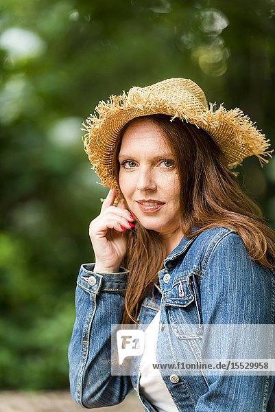 40 year old redheaded woman wearing a straw hat and a jean jacket looking at the camera  outdoors.