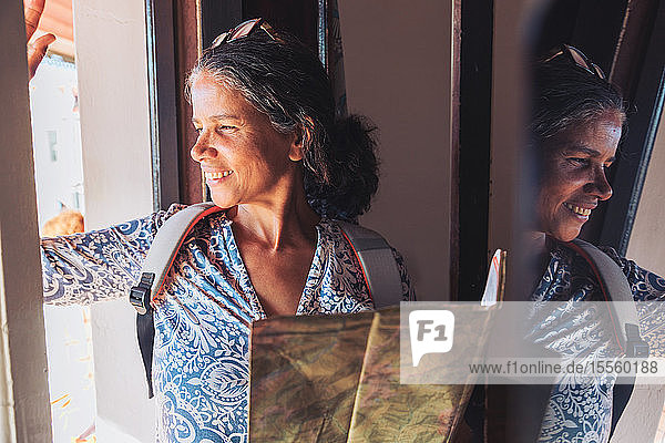 Lächelnde Touristin mit Landkarte schaut aus dem sonnigen Fenster