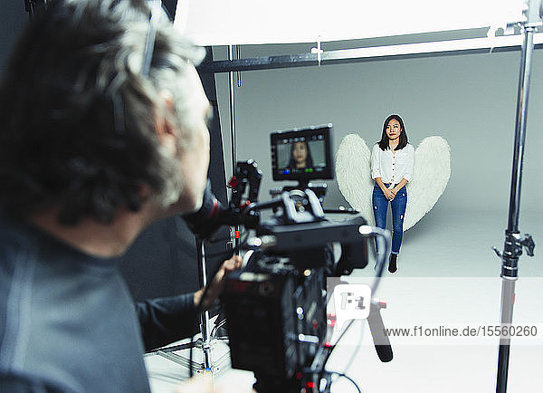 Photographer behind camera photographing young woman wearing angel wings during photo shoot in studio