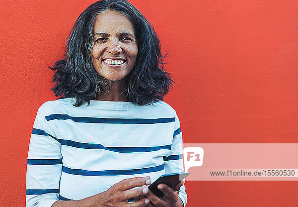 Porträt einer lächelnden  selbstbewussten Frau  die ein Smartphone vor rotem Hintergrund benutzt