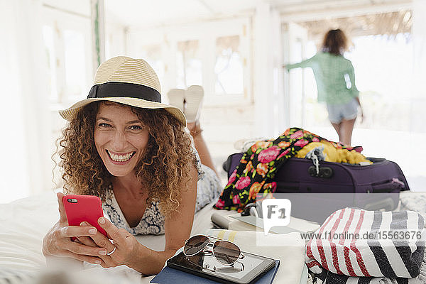 Porträt einer glücklichen jungen Frau  die ihr Smartphone benutzt und ihren Koffer im Schlafzimmer auspackt