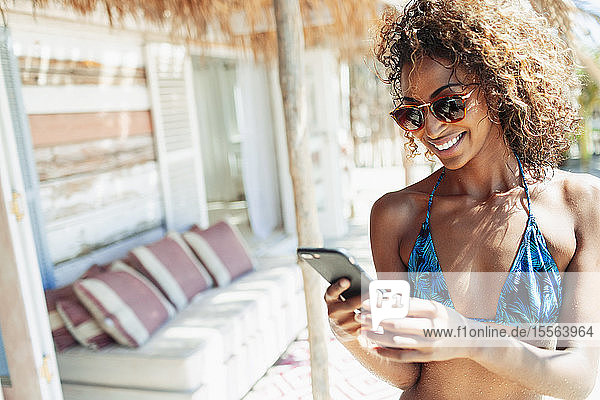 Glückliche junge Frau im Bikini  die ihr Smartphone auf der sonnigen Terrasse einer Strandhütte benutzt
