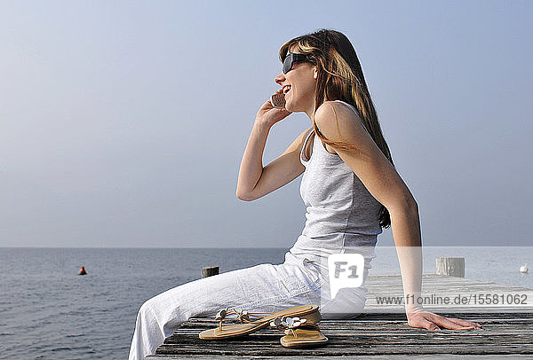 Italien,  Gardasee,  Womann 20-25) auf der Kante des Docks sitzend mit Mobiltelefon,  Seitenansicht