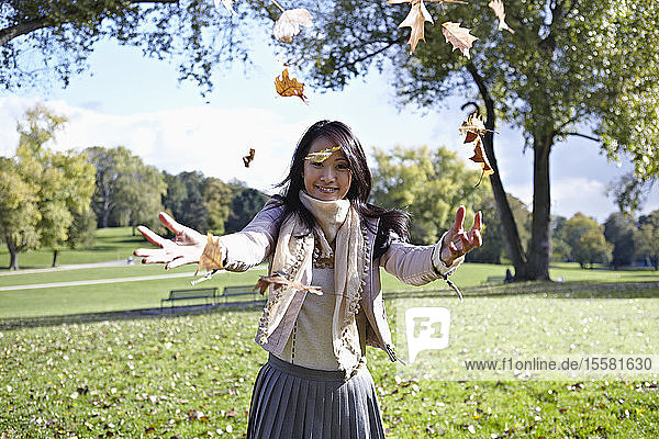 Deutschland  Köln  Junge Frau spielt im Park mit Blättern  lächelnd  Porträt