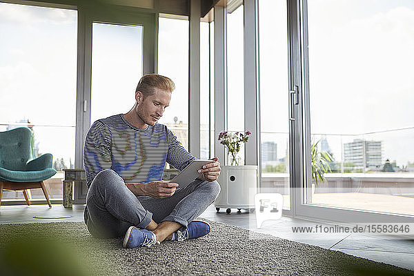 Junger Mann sitzt zu Hause mit Tablette auf Teppich