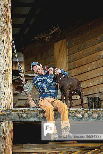 Mann streichelt Schokoladen-Labrador auf der Veranda einer Blockhütte