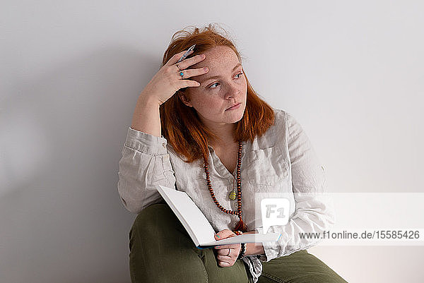 Junge Frau mit roten Haaren sitzt mit Tagebuch in der Hand und schaut zu Hause weg
