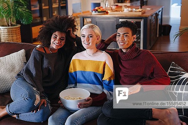 Junger Mann und junge Freundinnen sehen auf einem Wohnzimmersofa mit einer Schüssel Popcorn fern