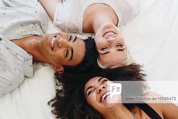 Junger Mann und zwei Freundinnen liegen lachend auf dem Bett  Blick von oben