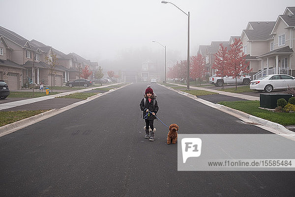 Mädchen geht mit dem Hund inmitten einer nebligen Vorstadtstraße spazieren  Portrait in voller Länge