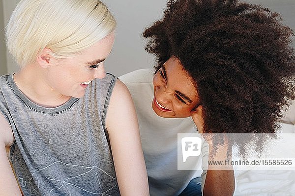 Two happy young female friends sitting on bed chatting