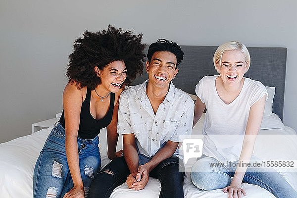 Young man and two female friends sitting on bed laughing
