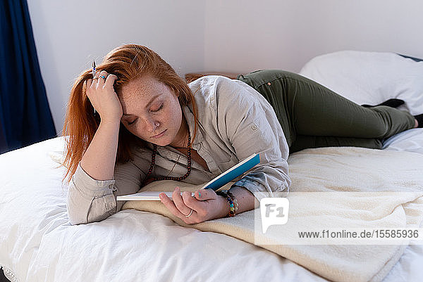 Junge Frau mit roten Haaren liegt auf dem Bett und liest ihr Tagebuch
