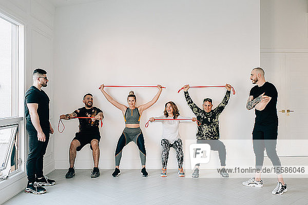 Fitnesstrainer beobachten Freunde  die im Studio mit Widerstandsband gegen die Wand hocken