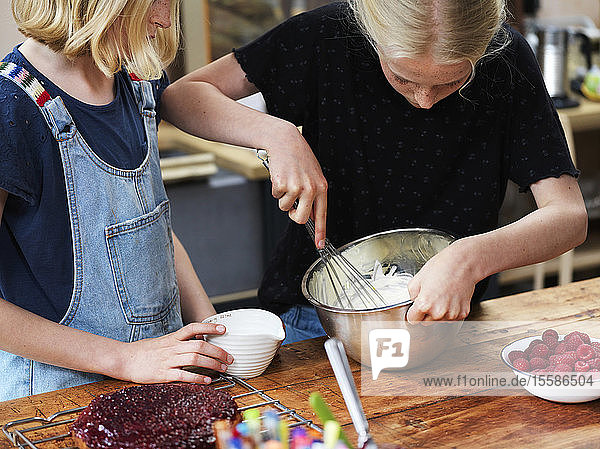 Mädchen und ihre Schwester backen einen Kuchen und verquirlen Sahne in Rührschüssel am Küchentisch