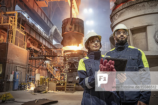 Male and female steelworkers using digital tablet during steel pour in steelworks
