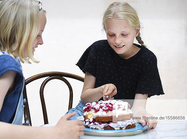 Mädchen und ihre Schwester schneiden ihren selbstgebackenen Kuchen mit frischen Himbeeren am Küchentisch an