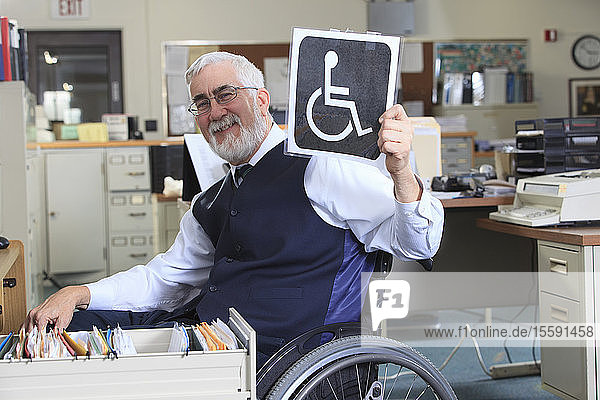 Man with Muscular Dystrophy in a wheelchair working in an office and holding up a Handicapped sign