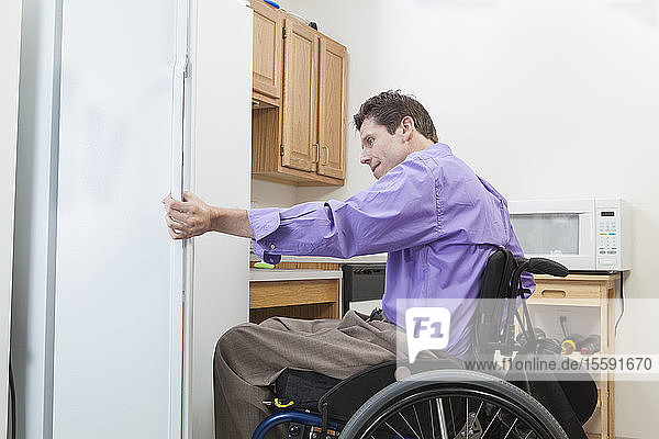 Mann im Rollstuhl mit Rückenmarksverletzung beim Öffnen eines Kühlschranks in einer barrierefreien Küche