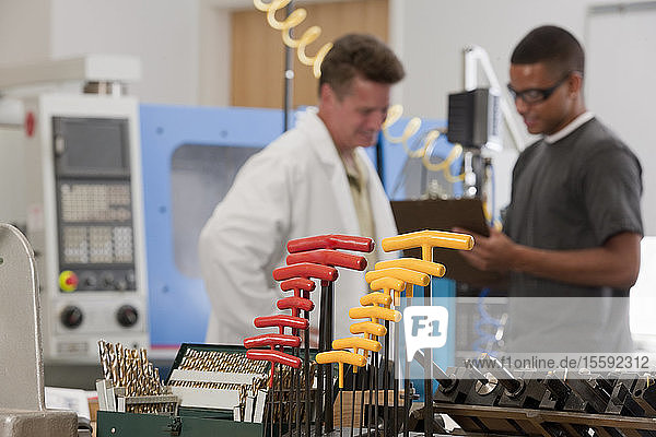 Ein Ingenieurprofessor und ein Student diskutieren über eine CNC-Maschine im Labor mit T-Griff-Schlüsseln und Bohrern im Vordergrund