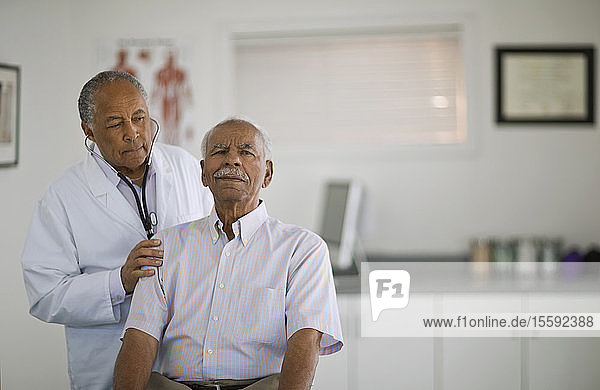 Ein älterer Mann lässt sich von einem männlichen Arzt in einem Büro den Herzschlag abhören.
