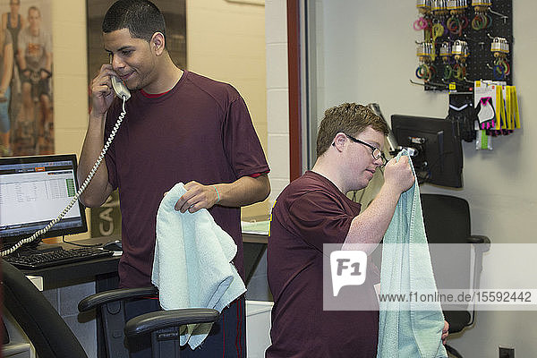 Young man with Down Syndrome preparing towels at college equipment dispensary for gym with supervisor using phone