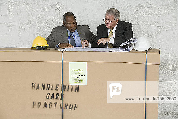 Zwei Geschäftsleute sitzen an einer Transportkiste und besprechen einen Bauplan