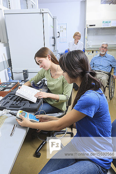 Ingenieurstudenten bei der Arbeit an einem chemischen Analysekit mit Professoren im Hintergrund  einer davon mit Muskeldystrophie