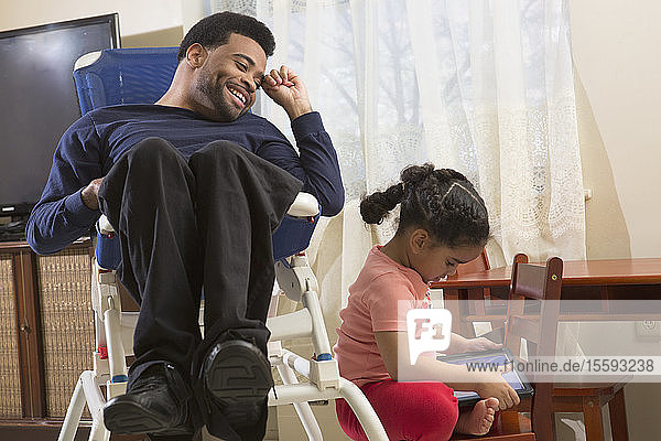 Glücklicher afroamerikanischer Mann mit zerebraler Lähmung und seiner Tochter  die zu Hause ein Tablet benutzt