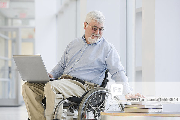 Universitätsprofessor mit Muskeldystrophie liest ein Buch und benutzt einen Laptop
