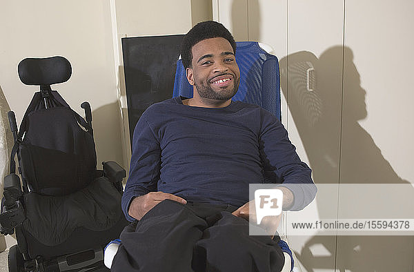 Glücklicher afroamerikanischer Mann mit Zerebralparese sitzt zu Hause neben seinem Rollstuhl